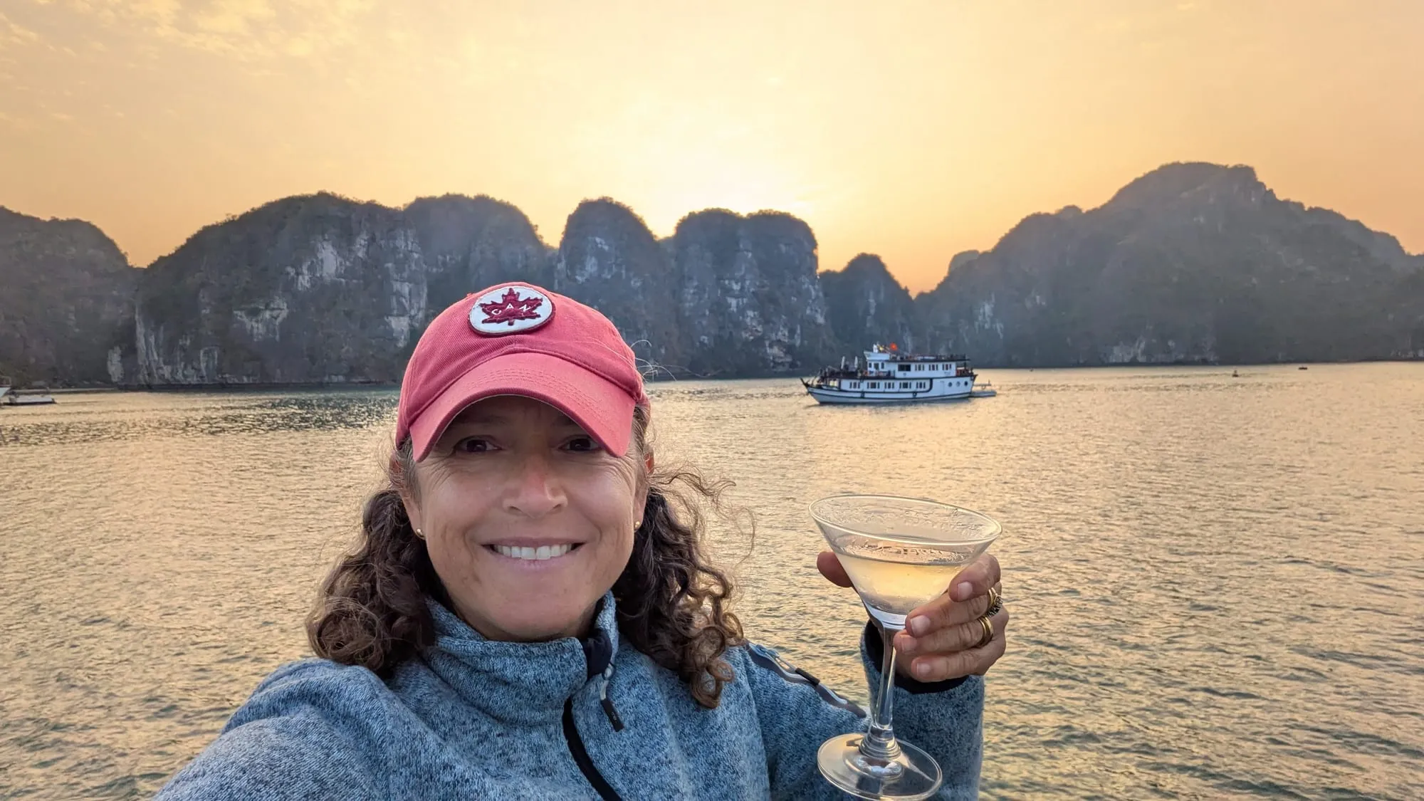 Kim raising a glass at sunset on Ha Long Bay, limestone karsts glowing behind her