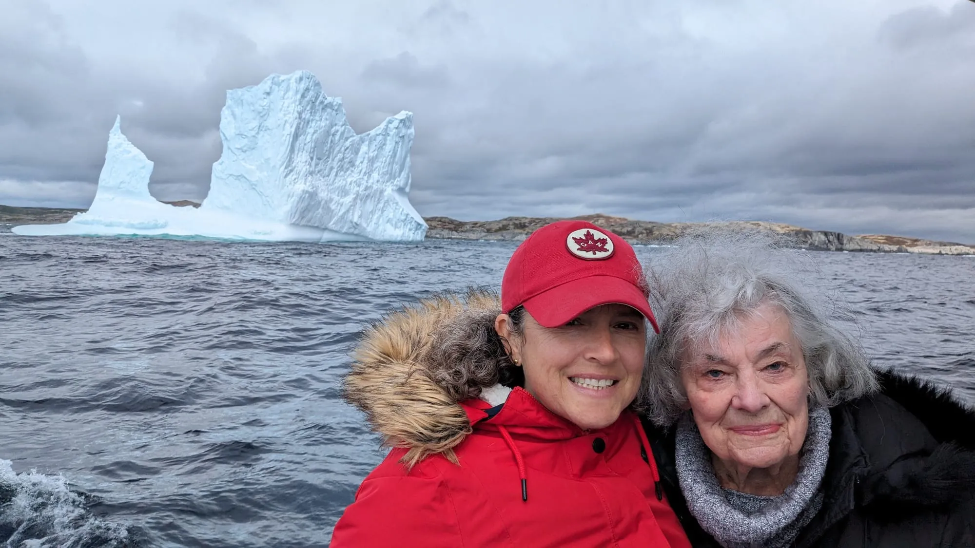 Kim and Dee bundled in red jackets on a boat, smiling in front of a towering iceberg