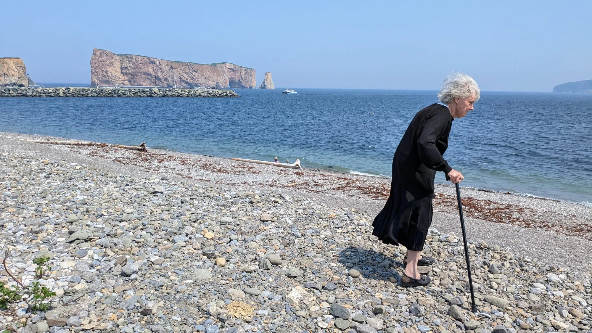 Dee walking with a cane along a pebble beach, with Percé Rock rising from the sea behind her